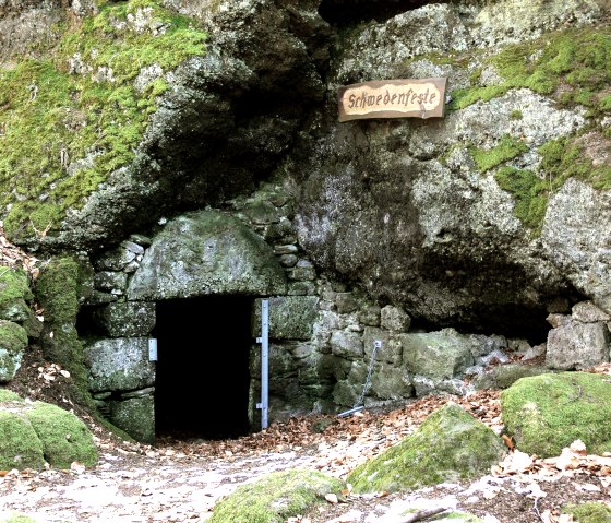 Een met mos bedekte rots met een donkere grotingang, met het opschrift 'Schwedenfeste'. Herfstbladeren bedekken de grond., © Touristik GmbH Gerolsteiner Land