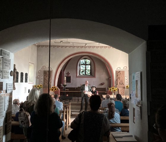 Mehrere Personen von hinten fotografiert sitzen in einer Kirche und schauen nach vorne, wo zwei Personen vorlesen.