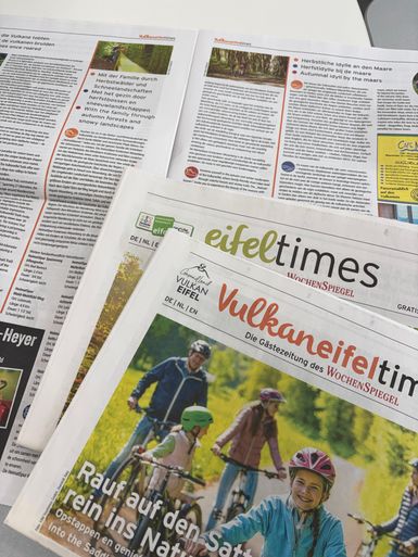 Aufgeschlagene Zeitungen mit Titeln "Eifeltimes" und "Vulkaneifeltimes" und Fotos von Familienurlaub.
