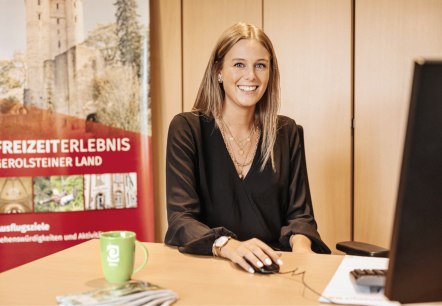 Eine Frau sitzt an einem Schreibtisch und lächelt in die Kamera. Hinter ihr einige Schränke und ein Banner mit Aufdruck &quot;Freizeirerlebnis Gerolsteiner Land&quot; und Bild einer Burg.