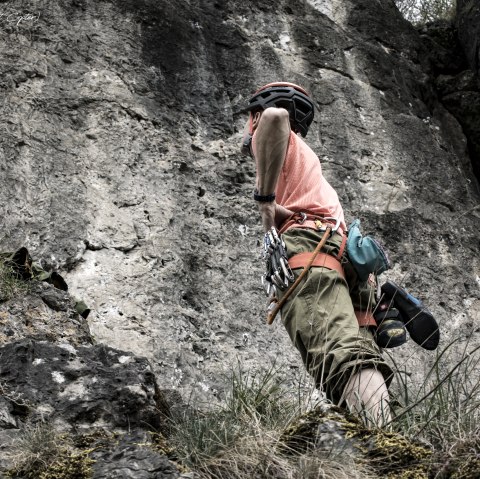 A person wearing a helmet and equipment stands in front of a steep rock face, hands on hips, looking up.