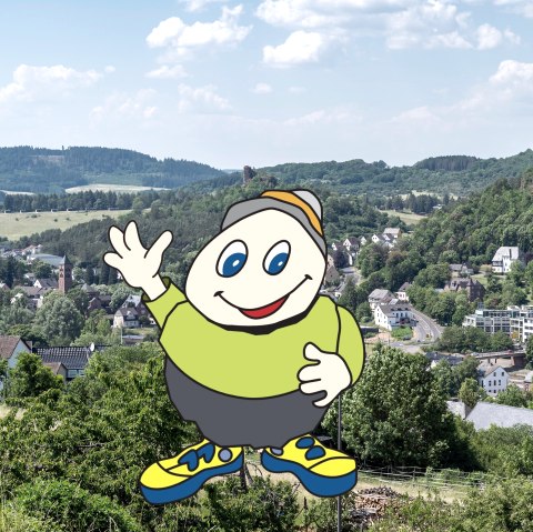 Cartoonfigur Willi Basalt mit grünem Shirt, grauer Hose und gelben Schuhen winkt. Hinter ihm Blick auf die Stadt Gerolstein mit Kirche, Gebäuden und Felsen.