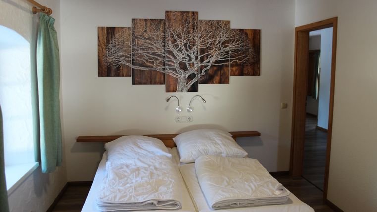 Ein gemütliches Schlafzimmer mit zwei Betten und einem dekorativen Wandbild eines Baumes. Die Wände sind hell und die Fenster haben grüne Vorhänge.