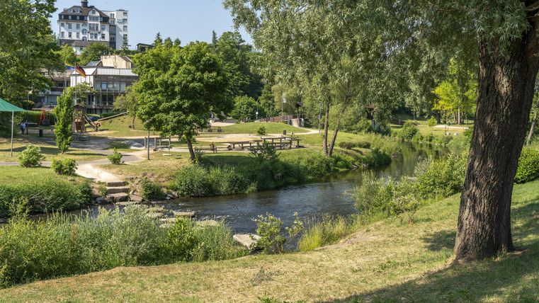 Ein grüner Park mit einem Fluss, Bäumen und einem Gebäude im Hintergrund. Es gibt Picknicktische und einen Spielplatz.