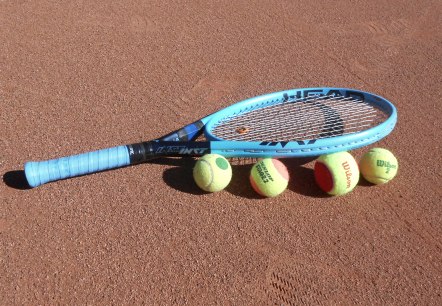 Ein  blauer Tennisschl&auml;ger liegt auf und neben 4 Tennisb&auml;llen auf einem Tennisplatz.