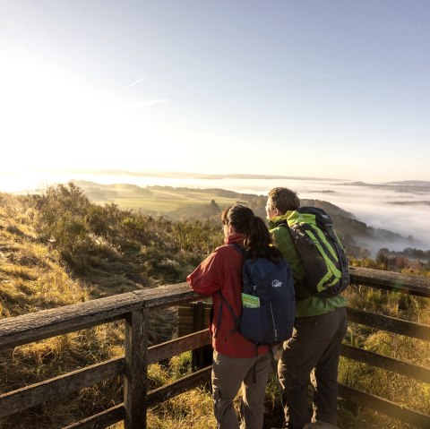 Zwei Wanderer stehen auf einer Aussichtsplattform auf dem Gipfel eiens Berges un schauen in die herbstliche und nebelige Landschaft.