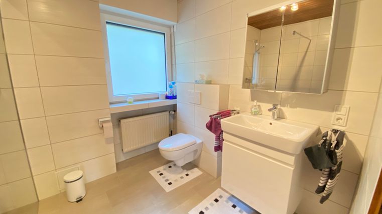 Ein modernes Badezimmer mit hellem Fliesenboden und weißen Wänden. Die Ausstattung umfasst ein Waschbecken, eine Toilette und ein Fenster, das Tageslicht hereinscheinen lässt.