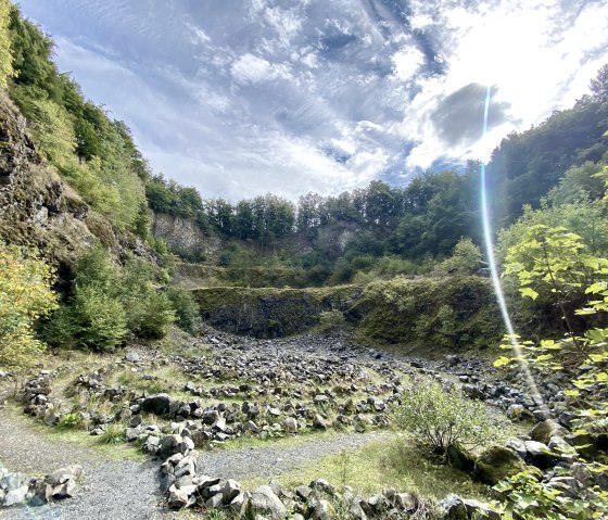 Interieur van de vulkaan Arensberg met stenige paden omgeven door groene vegetatie en rotsen onder een bewolkte hemel met zonnestralen., © Touristik GmbH Gerolsteiner Land