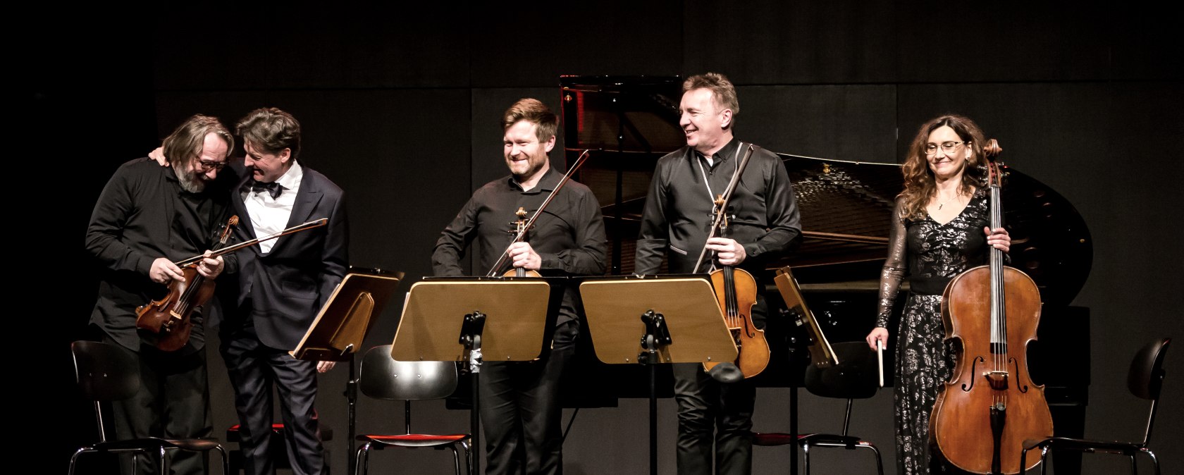 F&uuml;nf Musikanten auf einer B&uuml;hne mit Streichinstrumenten in der Hand stehen nebeneinander und lachen.