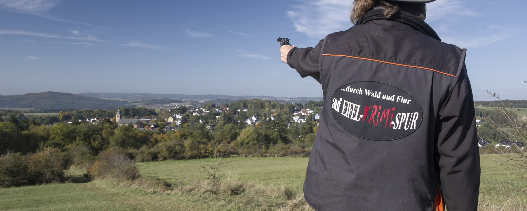Eine Hut tragende Person steht mit dem Rücken zum Betrachter und zeigt mit einer Pistole in die Ferne. Sie trägt eine Jacke mit der Aufschrift &quot;...durch Wald und Flur auf Eifel-Krimi-Spur&quot; auf dem Rücken. Vor der Person liegt eine große Wiese und dahinter in der Ferne ein Dorf mit  vielen Häusern.