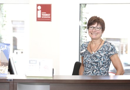 A woman stands behind a counter and smiles at the camera. Behind her are several windows and the i-mark logo for certified tourist information.