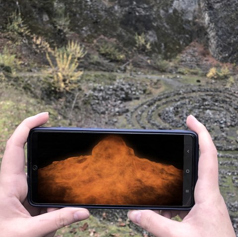 Two hands hold a smartphone across the abyss of a volcano. The volcanic explosion is visualised on the screen.