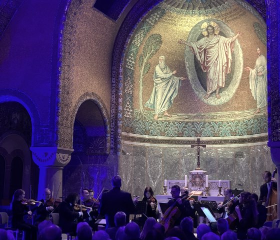 Schwach beleuchteter Innenraum der Erlöserkirche Gerolstein mit goldener Mosiakverzierung. Im Altarraum Musikanten und ein großes Publikum in den Sitzbänken.
