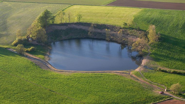 Vogelperspektive von oben auf den kreisrunden See des Eichholzmaares. Das Maar liegt mitten zwischen Wiesen und Feldern. Ein Fußweg führt um das Maar herum.
