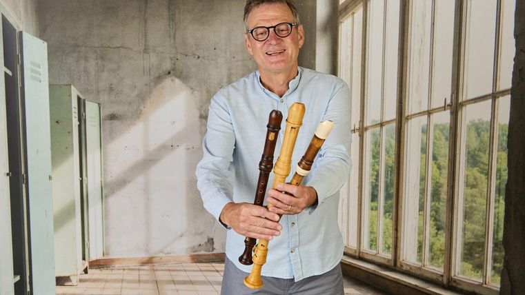 Mann steht im Vordergrund und hält drei verschiedene Flöten in der Hand, der Mann lächelt und schaut auf die Flöten, er steht in einem hell beleuchteten Raum