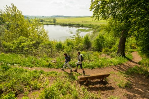 Zwei Wanderer auf einem Pfad am Eichholzmaar in der Eifel, umgeben von grüner Natur und einem See im Hintergrund.