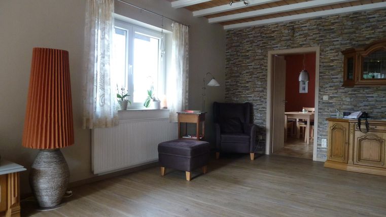 A cozy living room with a large window and wooden floor. There is a floor lamp, two armchairs, and a small table.