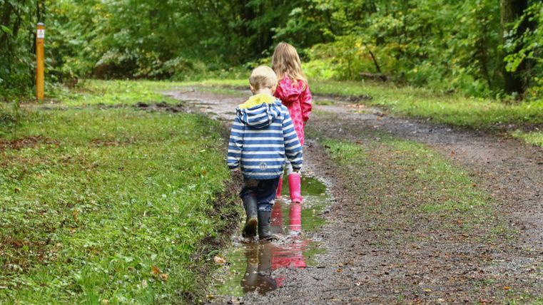 Zwei in Regenjacken und Gummistiefeln gekleidete Kinder laufen auf einem Waldpfad durch eine Pfütze.