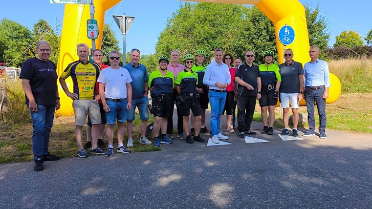 Zahlreiche Personen, welche teilsweise sportlicher Radfahrkleidung tragen, stehen auf dem Radweg unter einem großen Luftbogen des Sponsors e-regio.