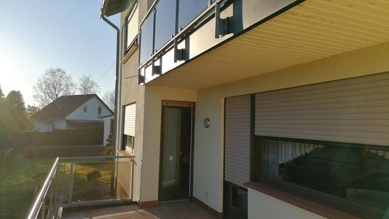 A modern balcony with glass railing in the sunlight. In the background, houses and a clear sky are visible.
