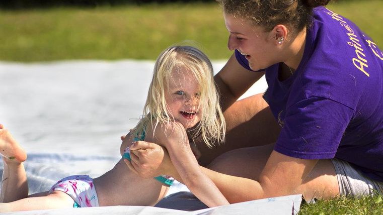 Ein fröhliches Kind mit blonden Haaren spielt auf einer Wiese. Eine Erwachsene sitzt neben ihr und hilft ihr beim Spielen.