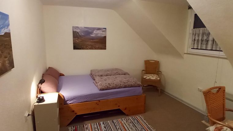 Ein gemütliches Zimmer mit einem Bett und einem kleinen Tisch. An den Wänden hängen Bilder, und ein Stuhl ist neben dem Bett platziert.