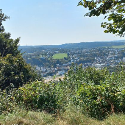 Eine Landschaft mit Blick auf eine Stadt, umgeben von grünen Hügeln. Der Himmel ist klar und blau.