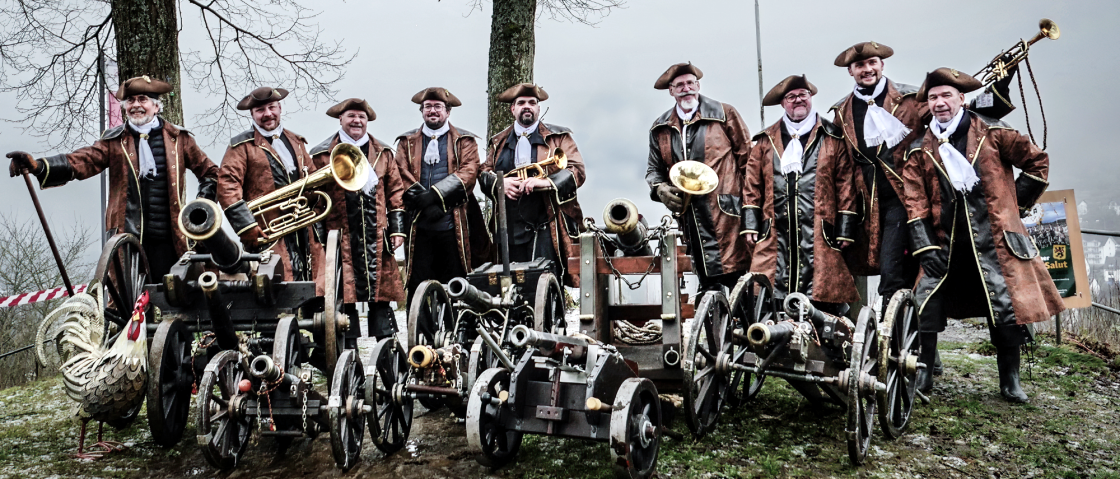 Auf einem Foto posiert eine Gruppe von M&auml;nnern in historischer Kleidung, umgeben von kleinen Kanonen und Musikinstrumenten, vor dem Hintergrund einer tr&uuml;ben, winterlichen Landschaft.Die Szene zeigt eine Gruppe von neun M&auml;nnern, die sich auf einer leicht erh&ouml;hten Grasfl&auml;che aufstellen, mit mehreren kleinen Kanonen vor sich und Musikinstrumenten wie Trompeten und einer Posaune. Sie scheinen entweder f&uuml;r ein Gruppenfoto zu posieren oder sich auf eine Vorf&uuml;hrung vorzubereiten. Die Kanonen sind in einer Reihe angeordnet, mit einem dekorativen metallenen Hahn auf der linken Seite. Im Hintergrund ist eine bew&ouml;lkte Landschaft mit B&auml;umen und einem H&uuml;gel mit einer Stadtansicht zu sehen, sowie eine Fahne mit einem Wappen. Die Komposition ist symmetrisch, wobei die M&auml;nner und Kanonen eine dominante horizontale Linie bilden, die durch die vertikalen Elemente der B&auml;ume und die Fahne ausgeglichen wird.Die Hauptdarsteller sind neun M&auml;nner unterschiedlichen Alters, die traditionelle Kleidung aus dem 18. Jahrhundert tragen. Sie alle sind in braunen R&ouml;cken mit schwarzen Jacken, wei&szlig;en Hemden und Halsbinden sowie Dreispitzenh&uuml;ten gekleidet. Einige halten Musikinstrumente wie Trompeten und eine Posaune, w&auml;hrend andere die H&auml;nde an ihren H&uuml;ften oder auf einem Gehstock ruhen lassen. Ihre Gesichtsausdr&uuml;cke variieren, einige l&auml;cheln, w&auml;hrend andere ernster blicken, was der Szene eine gewisse Lebendigkeit verleiht. Die Kanonen, die sie umgeben, sind detailreich gestaltet und aufwendig verziert, was auf ihre historische Bedeutung hinweist.Die fotografische Ausf&uuml;hrung wirkt professionell, mit einem klaren Fokus auf die Hauptdarsteller und die Kanonen. Die Farbgebung ist nat&uuml;rlich und die Details sind gut erkennbar, was die Textur der Kleidung, der Kanonen und der Umgebung hervorhebt. Das Licht scheint von vorne zu kommen, was die Gesichter der M&auml;nner und die Details der Kanonen gut ausleuchtet. Der Stil der Fotografie ist dokumentarisch, was darauf hindeutet, dass das Foto wahrscheinlich bei einer Veranstaltung oder einem historischen Reenactment aufgenommen wurde.Die Szene spielt sich an einem bew&ouml;lkten Tag ab, was eine eher ged&auml;mpfte und k&uuml;hle Atmosph&auml;re erzeugt. Der Hintergrund ist verschwommen, was die Hauptdarsteller hervorhebt, w&auml;hrend die B&auml;ume und die Stadtansicht im Hintergrund einen Kontext f&uuml;r die Szene schaffen. Der Boden ist mit nassem Gras bedeckt, was auf die Jahreszeit hindeutet und die raue Natur der Umgebung verst&auml;rkt. Die leicht getr&uuml;bte Beleuchtung und der Hintergrund schaffen eine ged&auml;mpfte, realistische Atmosph&auml;re, die gut zur historischen Thematik passt., &copy; Klaus Wollwert 