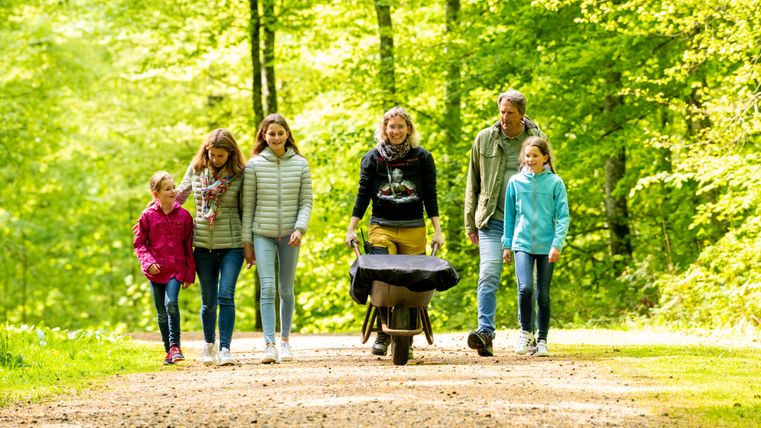 Sechs Personen, drunter drei Kinder, laufen nebeneinander auf einem Waldpfad durch einen strahlend grünen Wald. Eine der Frauen schiebt eine Schubkarre.