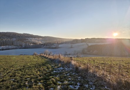 H&uuml;gelige Wiesenlandschaft leicht mit Schnee bedeckt und Sonnenuntergang im Hintergrund.