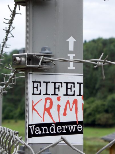 Ein Schild mit der Aufschrift 'Eifel Krimi Wanderweg' an einem Stacheldrahtzaun.