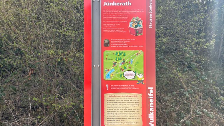 Eine Informationstafel des Märchenpfads Jünkerath in der Vulkaneifel, umgeben von Büschen.