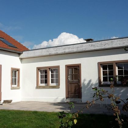 Een modern, helder huis met witte muren en houten ramen. De lucht is blauw en er zijn enkele wolken zichtbaar.