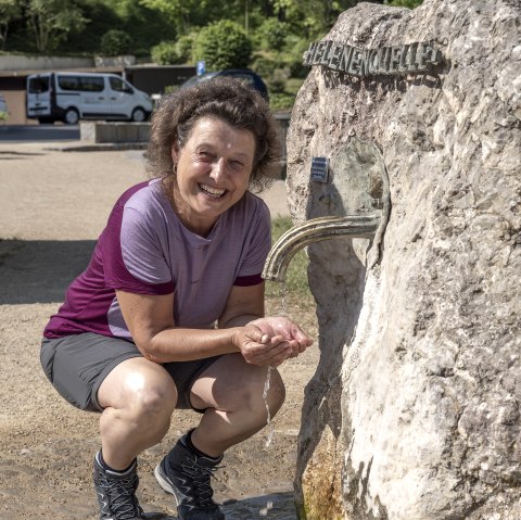 Eine Frau kniet neben einem Felsen aus dem ein Hahn, aus welchem Wasser herausstr&ouml;mt. Die Frau f&auml;ngt das Wasser mit den H&auml;nden auf.