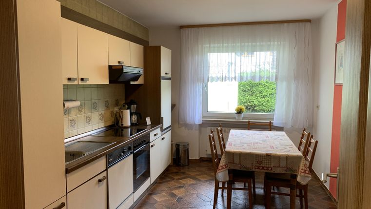 Eine helle Küche mit modernen Geräten und einem Essbereich. Ein Tisch mit Stühlen steht unter einem großen Fenster.