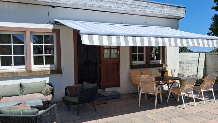 Eine gemütliche Terrasse mit einem Markisen und verschiedenen Sitzgelegenheiten. Der Bereich ist hell und einladend, ideal zum Entspannen im Freien.