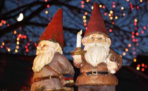Two decorative garden gnomes, one of whom is holding a candle. Colorful lights are glowing in the background.
