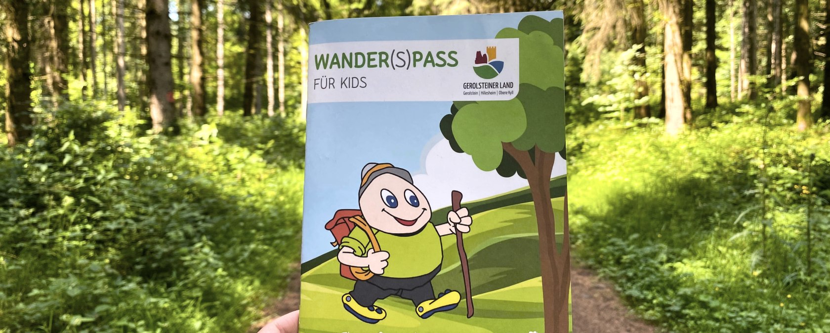 Das Prospekt "Wander(S)pass f&uuml;r Kids" wird hochgehalten mit einem dichten Wald und Waldweg im Hintergrund.
