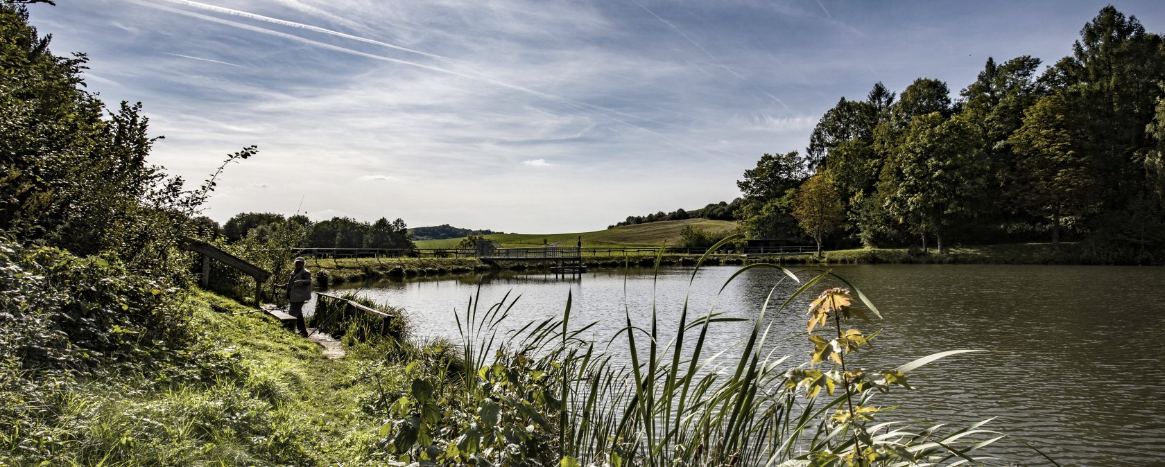 Ein ruhiger Stausee mit gr&uuml;nem Ufer, B&auml;umen und einem klaren Himmel mit Kondensstreifen. Eine Person steht am Ufer., &copy; Martin M&uuml;ller