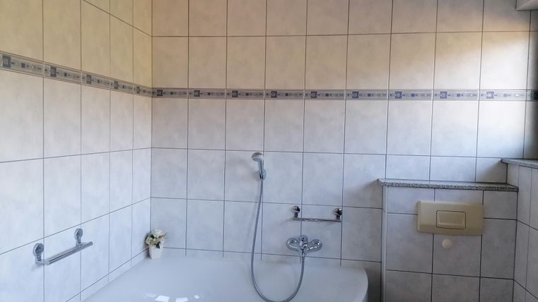 A modern bathroom with a white bathtub and a shower. The walls are tiled and there is a toilet and a towel holder.