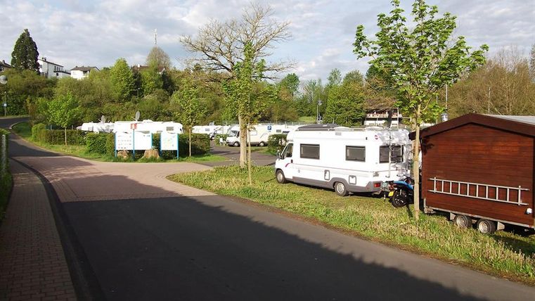 Wohnmobilstellplatz mit mehreren Wohnmobilen, Bäumen und einem kleinen Holzschuppen an einer asphaltierten Straße.