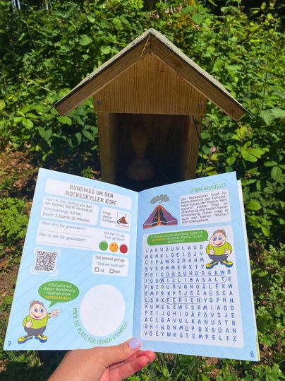 Ein geöffneter Wanderpass mit Informationen und einem Suchrätsel, gehalten vor einem kleinen Holzhaus im Grünen.