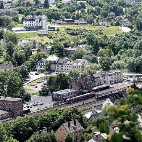 View from above of parts of Gerolstein with the railway station, where a train is currently stopping. Surrounded by trees, car parks and numerous buildings.