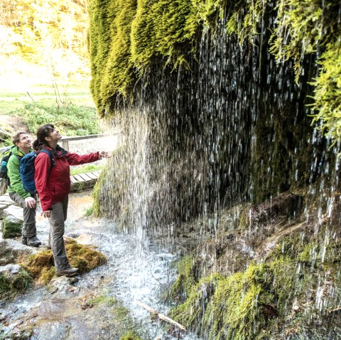 Zwei Personen stehen vor dem moosbedeckten Wasserfall. Sie tragen Outdoor-Kleidung und schauen fasziniert auf das herabfließende Wasser., © Eifel Tourismus GmbH, Dominik Ketz
