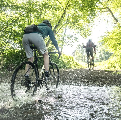 A cyclist on a gravel bike rides through the middle of a large fountain in a forest. The water splashes on all sides. Another cyclist rides out of the forest in front of him.