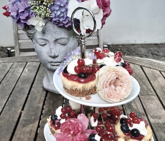 Etagere with small cakes, decorated with berries and flowers, on a wooden table. In the background, a flower vase in the shape of a head with colorful flowers., © Touristik GmbH Gerolsteiner Land, Ute Klinkhammer