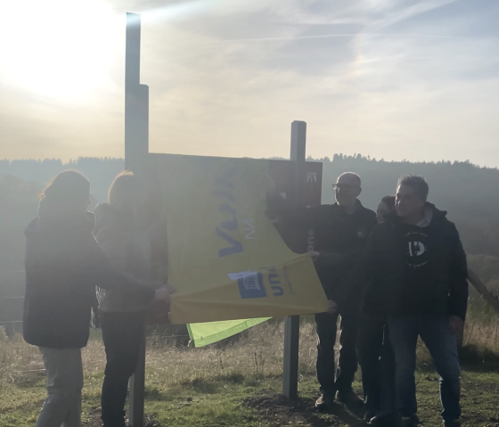 Fünf Personen stehen neben einem verhüllten Informationsschild und enthüllen es. Sonnenuntergang im Hintergrund.