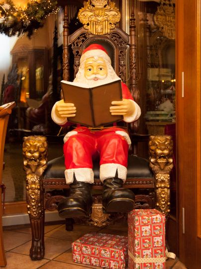 Ein weihnachtlicher Weihnachtsmann sitzt auf einem prächtigen Stuhl und liest ein Buch. Unter ihm liegen festlich verpackte Geschenke.