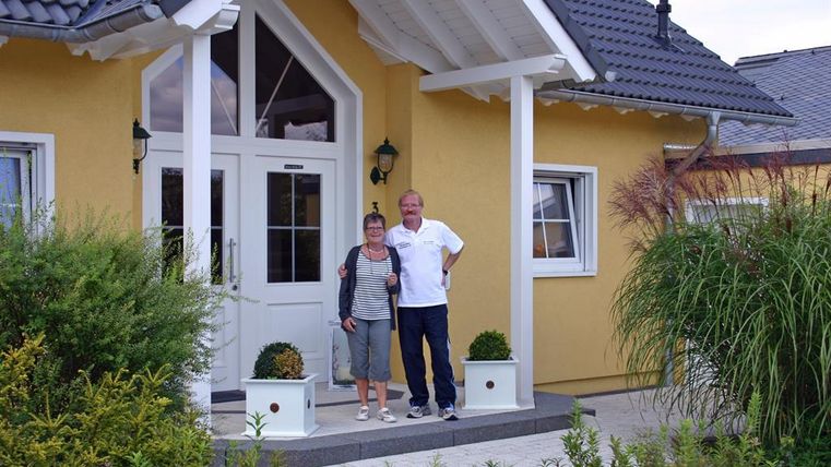 Een paar staat voor een geel huis met een brede ingangsv dávn. Omgeven door groene planten en een gastvrije sfeer.