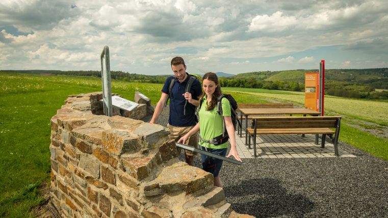 Zwei Wanderer betrachten eine Informationstafel an einer römischen Villa-Ruine auf einem Wanderweg.